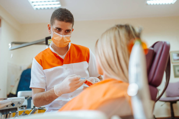 Μια όμορφη λευκή γυναίκα κάθεται σε μια καρέκλα στο γραφείο του οδοντιάτρου ενώ ο νεαρός οδοντίατρος εξετάζει τα δόντια της. - Φωτογραφία, εικόνα