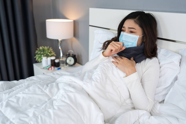 sick woman in medical mask coughing and suffering from virus disease and fever in a bed, coronavirus (covid-19) pandemic concept - 写真・画像