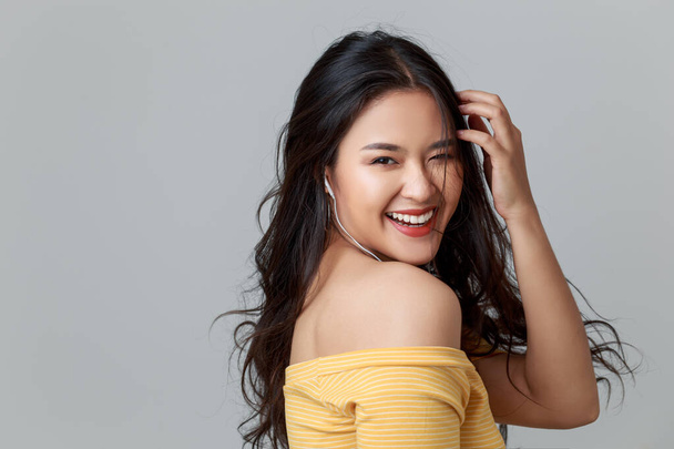 Close up jovem atraente mulher asiática vestindo camiseta amarela, fones de ouvido ouvindo música com rosto sorridente, estúdio cabeça tiro isolado no fundo cinza
. - Foto, Imagem