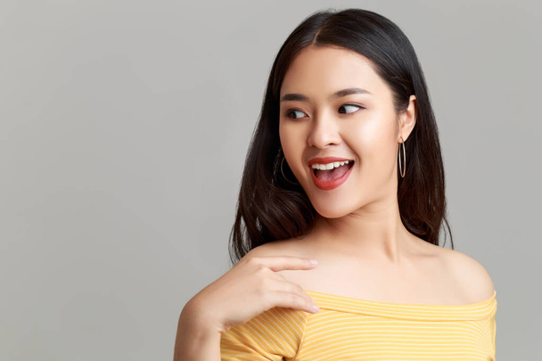 Close up jovem feliz atraente mulher asiática vestindo amarelo mostrando vestido de ombro com rosto sorridente doce isolado no fundo cinza
. - Foto, Imagem