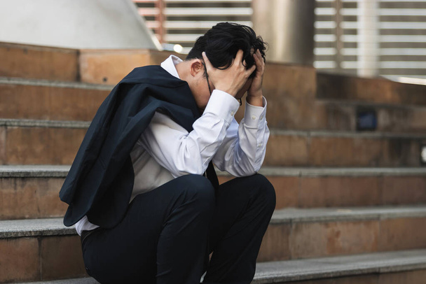 Aufgebracht sitzt ein junger asiatischer Geschäftsmann im Anzug mit den Händen auf dem Kopf auf der Treppe. Arbeitslosen- und Entlassungskonzept. - Foto, Bild