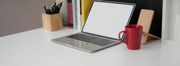 Captura recortada de mesa de trabajo elegante con computadora portátil de pantalla en blanco, taza, papelería y libros en escritorio blanco
   - Foto, Imagen