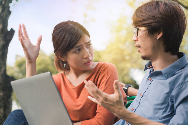 Young business asian man and woman working with them laptop and talking serious  on a bench in the park outdoors on vacation time. - 写真・画像