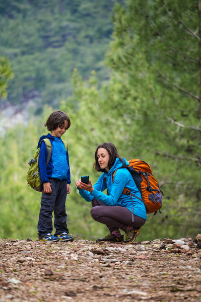 Mère montre à son enfant comment utiliser un smartphone pour localiser dans la forêt, garçon regarde un téléphone portable, Une femme avec un sac à dos voyage avec son fils dans les montagnes, Randonnée avec des enfants, géolocalisation, selfie
. - Photo, image
