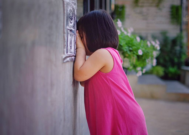 Мила азіатська дівчина з рожевим платтям грає в шкуру і шукає, рахуючи цифри з головою до бетонної стіни..  - Фото, зображення