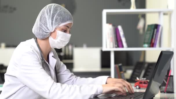 Kobieta w masce pracuje nad czymś z komputerem i odnotowuje to w biurze lub szpitalu..  - Materiał filmowy, wideo