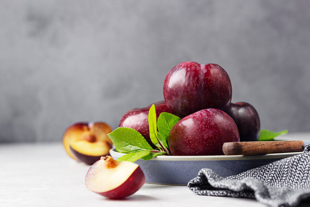 Whole purple plums and slices with leaves and knife on ceramic plate, light grey background. - Fotoğraf, Görsel