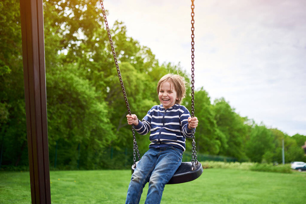 Outdoors portrait of cute preschool laughing boy swinging on a swing at the playground. - Fotografie, Obrázek