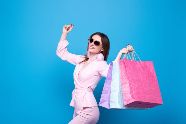 A fashionably dressed girl in sunglasses with multi-colored shopping bags is happy with the discounts and sale. Isolated on a blue background. - Fotoğraf, Görsel