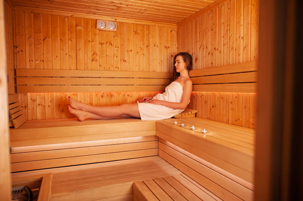 Die junge schöne Frau erholt sich in der Sauna. Konzeption der Körperpflege. Junge Frau sitzt auf Holzbank in Sauna. - Foto, Bild