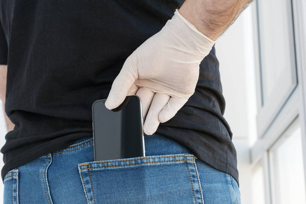  mužská ruka v bílé lékařské rukavici vytáhne černý smartphone ze zadní kapsy džín, ochranná a preventivní opatření proti bakteriím, virům, koronaviru, konceptu zdravotní péče, nový formát komunikace - Fotografie, Obrázek