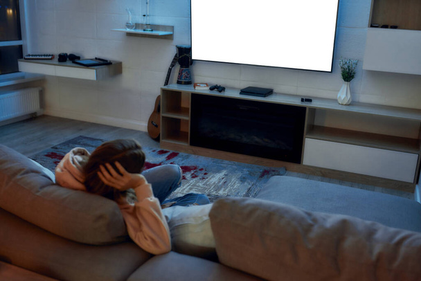 Evening vibes. Back view of woman sitting alone on the couch in front of TV set in the evening. Leisure and entertainment concept - Fotografie, Obrázek