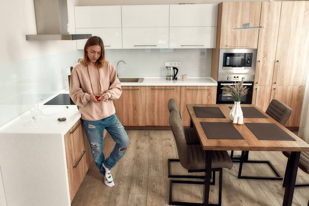 Getting ready. Young woman using red marijuana grinder, standing in the kitchen. Glass water pipe or bong, cannabis buds in a plastic bag on the table. Cannabis and weed legalization concept - 写真・画像