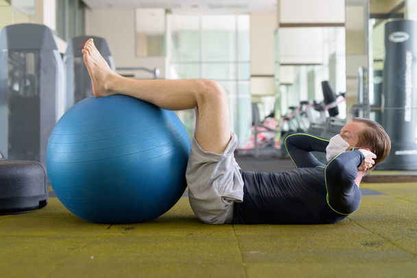 Full body shot of young man with mask doing sit ups with exercise ball at gym during corona virus covid-19 - Фото, изображение