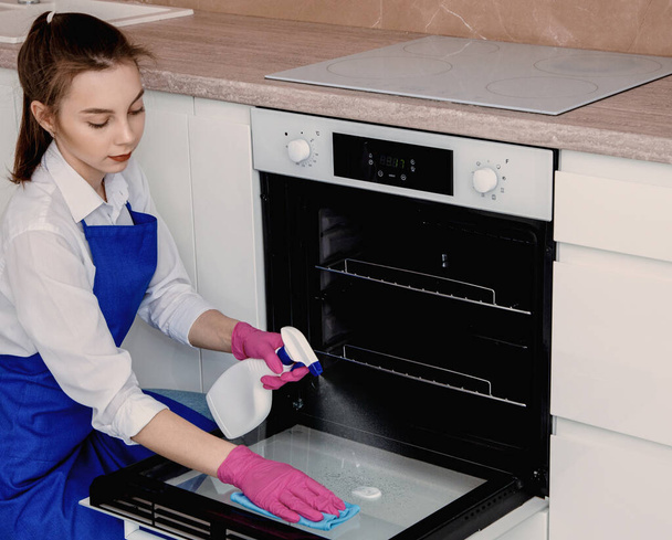 A girl washes an oven in a bright white kitchen with a spray in a white jar and a microfiber rag. - Foto, Imagen