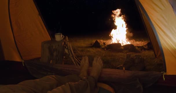 Turista v noci ve stanu ohřívá unavené nohy před krbem. Pohled zevnitř. Na obzoru - hvězdná obloha. - Záběry, video