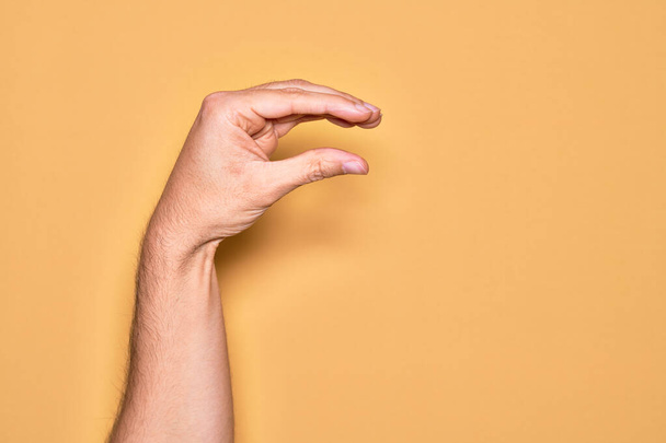 Χέρι του Καυκάσου νεαρός άνδρας δείχνει τα δάχτυλα πάνω από απομονωμένο κίτρινο φόντο συλλογή και τη λήψη αόρατο πράγμα, κρατώντας αντικείμενο με τα δάχτυλα που δείχνουν χώρο - Φωτογραφία, εικόνα