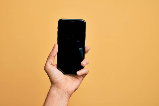 Hand of caucasian young man holding smartphone showing screen over isolated yellow background - Foto, Bild