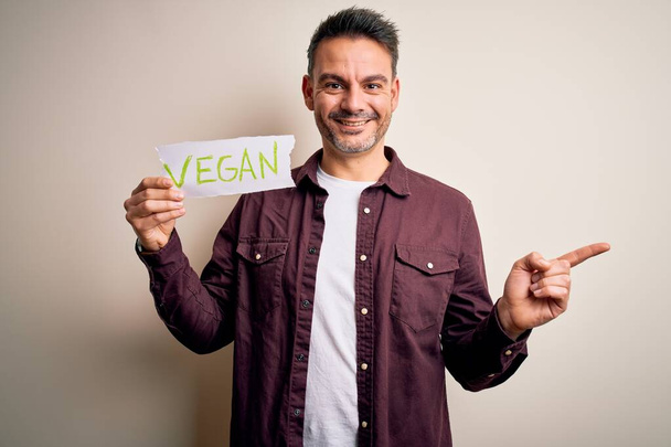 Jonge knappe veggie man met papier met veganistische boodschap over witte achtergrond zeer gelukkig wijzend met hand en vinger naar de zijkant - Foto, afbeelding