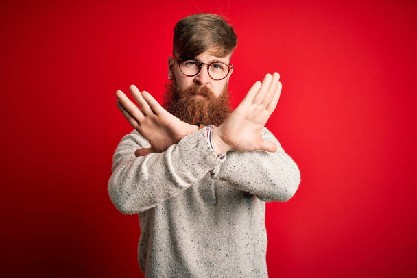 Handsome Irish redhead man with beard wearing casual sweater and glasses over red background Rejection expression crossing arms doing negative sign, angry face - Valokuva, kuva