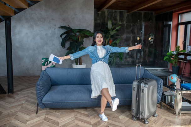 Dark-haired female, smiling, holding airline tickets, flopping down on sofa - Foto, afbeelding