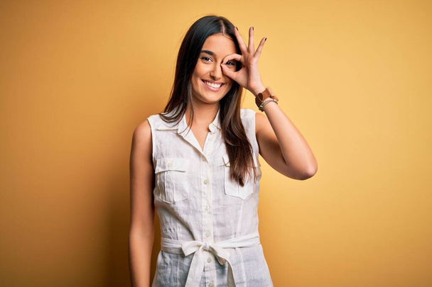 Young beautiful brunette woman on vacation wearing casual dress over yellow background doing ok gesture with hand smiling, eye looking through fingers with happy face. - Zdjęcie, obraz