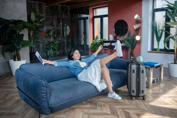 Dark-haired female, smiling, holding airline tickets, resting on sofa - Foto, Imagen