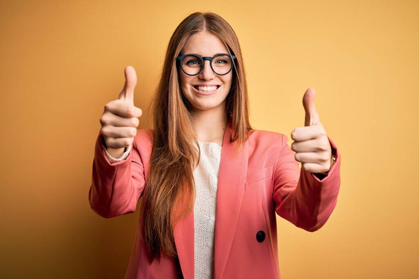 Young beautiful redhead woman wearing jacket and glasses over isolated yellow background approving doing positive gesture with hand, thumbs up smiling and happy for success. Winner gesture. - Photo, image