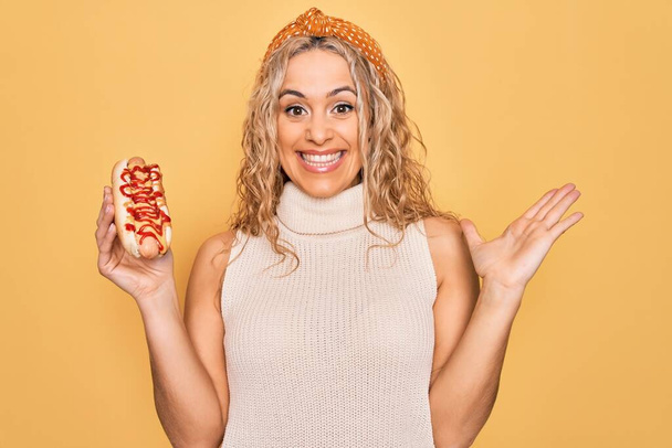 Молода красива блондинка їсть швидку їжу хот-дог з кетчупом і гірчицею, святкуючи перемогу з щасливою посмішкою і виразом переможця з піднятими руками
 - Фото, зображення