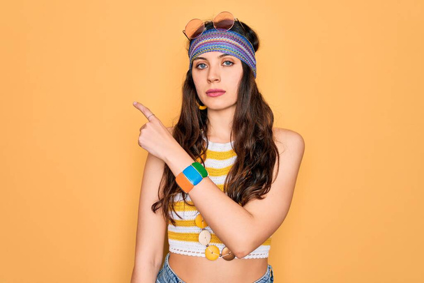 Fiatal gyönyörű hippi nő kék szemek visel kiegészítők és napszemüveg mutató kézzel ujját oldalra mutató reklám, komoly és nyugodt arc - Fotó, kép
