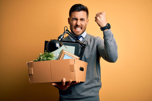 Young business man holding office box being fired from job over yellow background annoyed and frustrated shouting with anger, crazy and yelling with raised hand, anger concept - Photo, image