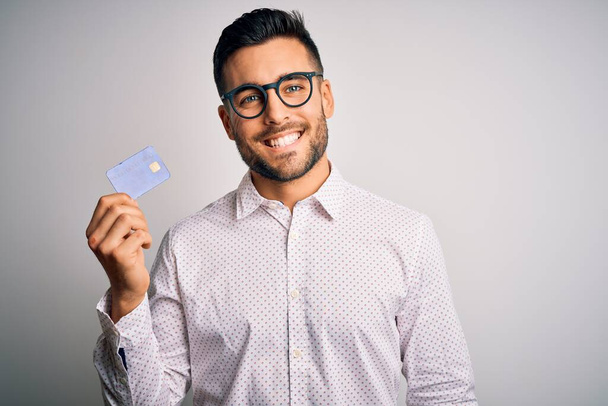 Joven hombre de negocios sosteniendo la tarjeta de crédito sobre un fondo aislado con una cara feliz de pie y sonriendo con una sonrisa segura que muestra los dientes
 - Foto, Imagen