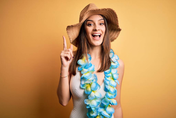 Jeune belle femme brune en vacances portant un maillot de bain et des fleurs hawaïennes lei pointant du doigt avec une idée réussie. Sorti et heureux. Numéro un
. - Photo, image
