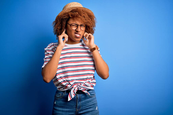 Афроамериканка с вьющимися волосами на отдыхе в летней шляпе и полосатой футболке, покрывающей уши пальцами с раздраженным выражением для шума громкой музыки. Глухая концепция
. - Фото, изображение