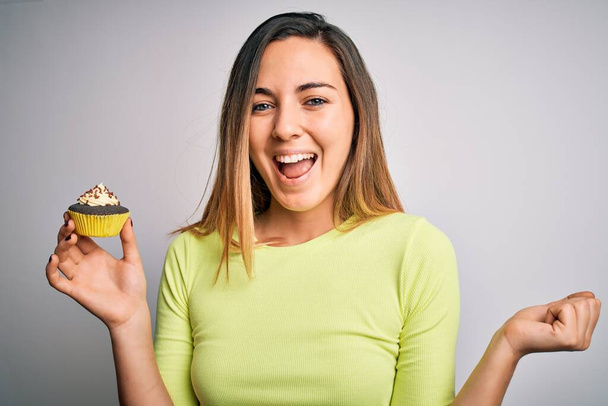 Mavi gözlü genç bir kadın beyaz arka planda tatlı çikolatalı kek yerken gururla bağırıyor, zaferi ve başarıyı kutluyor. Çok heyecanlı, coşkulu bir duygu. - Fotoğraf, Görsel