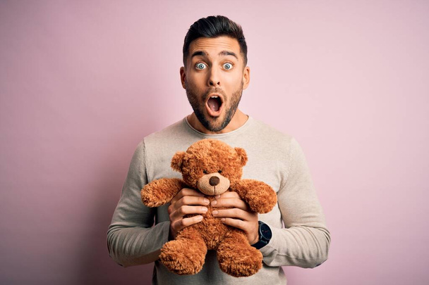 Jeune bel homme tenant un ours en peluche debout sur un fond rose isolé effrayé par un visage surpris, effrayé et excité par l'expression de la peur
 - Photo, image