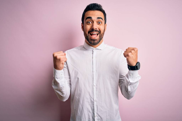 Young handsome man with beard wearing casual shirt standing over pink background celebrating surprised and amazed for success with arms raised and open eyes. Winner concept. - Foto, Bild