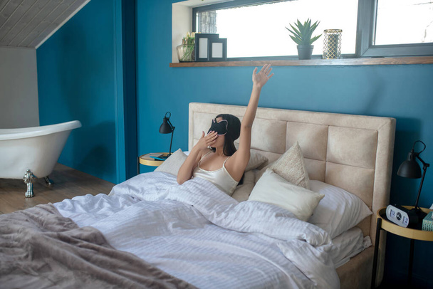 Young brunette lying in bed in sleep mask, stretching, yawning - Fotoğraf, Görsel