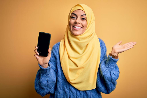 Joven hermosa mujer árabe morena con hiyab islámico sosteniendo teléfono inteligente mostrando la pantalla muy feliz y emocionada, la expresión ganadora celebrando la victoria gritando con una gran sonrisa y las manos levantadas
 - Foto, imagen