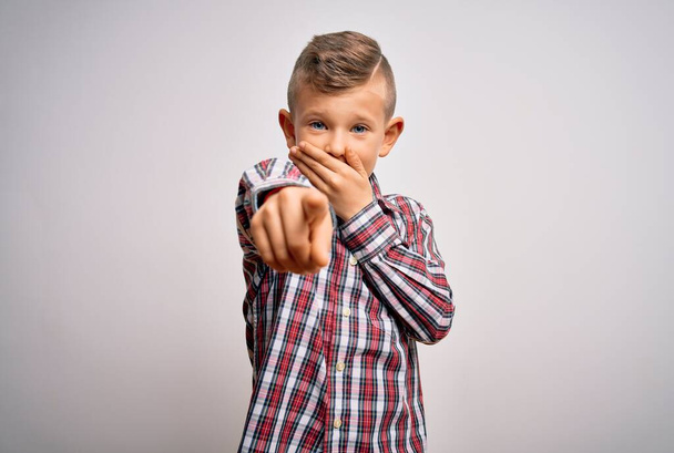 Nuori kaukasialainen sinisilmäinen poika yllään tyylikäs paita seisoo eristetyn taustan päällä nauraen sinulle, osoittaen sormella kameraa kädestä suuhun, häpeä ilme
 - Valokuva, kuva