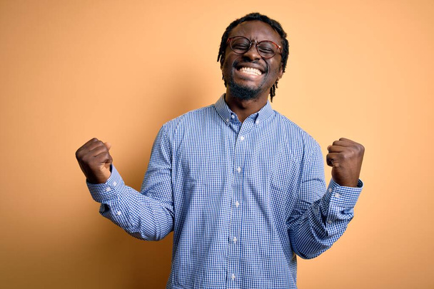 Młody, przystojny, afrykański mężczyzna w koszuli i okularach na żółtym tle bardzo szczęśliwy i podekscytowany wykonując gest zwycięzcy z podniesionymi ramionami, uśmiechnięty i wołający o sukces. Koncepcja uroczystości. - Zdjęcie, obraz