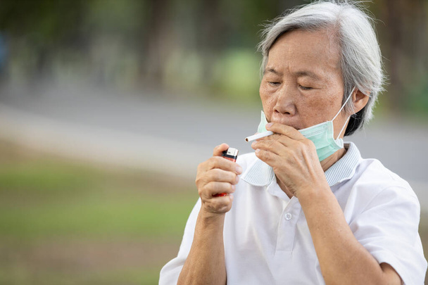 Oudere vrouw houdt een sigaret in de mond, ontwenningsverschijnselen, hunkeren naar sigaretten, zich gestrest voelen terwijl ze een masker draagt tijdens de pandemie Covid-19, Coronavirus, oudere vrouw is verslaafd aan roken - Foto, afbeelding