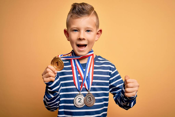 黄色の背景に賞のメダルを身に着けている若い白人の勝者の子供は誇りを持って叫び、勝利と成功を祝う非常に興奮し、応援感情 - 写真・画像