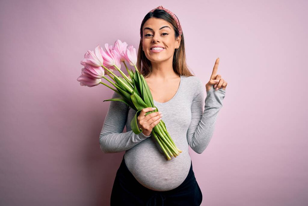 Молода красива брюнетка вагітна, очікуючи, що дитина тримає букет тюльпанів, здивована ідеєю або питанням, що вказує пальцем на щасливе обличчя, номер один
 - Фото, зображення