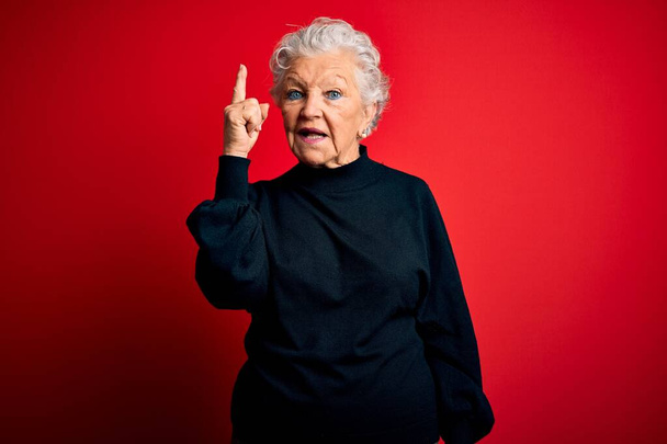 Senior beautiful woman wearing casual sweater standing over isolated red background pointing finger up with successful idea. Exited and happy. Number one. - Valokuva, kuva