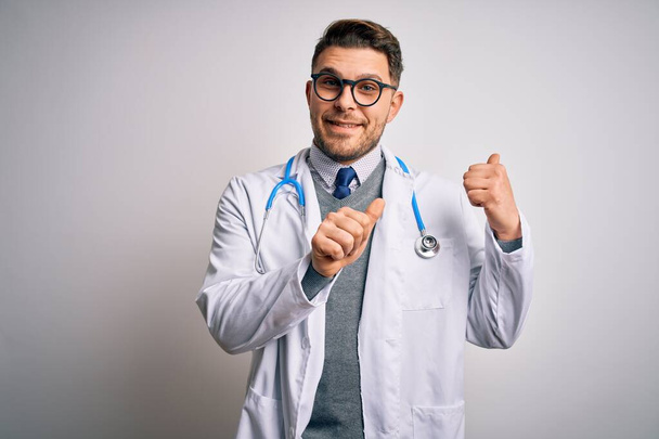 Młody lekarz z niebieskimi oczami, noszący płaszcz medyczny i stetoskop na odosobnionym tle, wskazujący na plecy z rękoma i kciukami w górze, uśmiechnięty pewny siebie - Zdjęcie, obraz