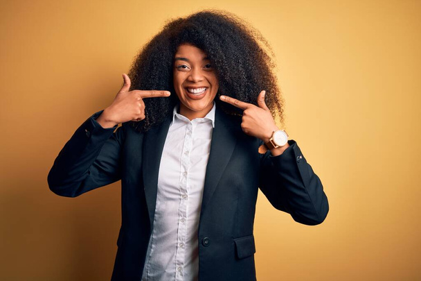 Jonge mooie Afrikaanse Amerikaanse zakenvrouw met afro haar dragen elegante jas lachend vrolijke tonen en wijzen met vingers tanden en mond. Tandheelkundige gezondheidszorg. - Foto, afbeelding