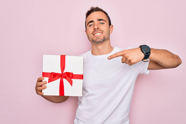 Young handsome man with blue eyes holding birthday present over isolated pink background very happy pointing with hand and finger - Фото, зображення