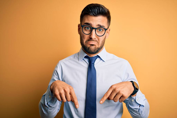 Young handsome businessman wearing tie and glasses standing over yellow background Pointing down looking sad and upset, indicating direction with fingers, unhappy and depressed. - Photo, Image