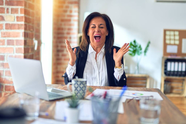 腕を上げて成功のために狂気と狂気を祝うオフィスでラップトップを使用して働いている中年の美しいビジネスマンの女性は興奮叫んで目を閉じています。勝者のコンセプト - 写真・画像